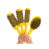 Four yellow hair styling brushes held by hands against a plain white background. Three are round barrel brushes and one is an oval paddle brush, all with dark bristles.